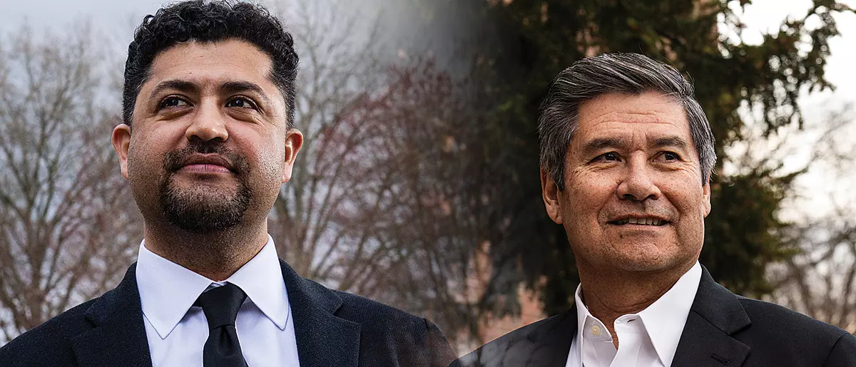 Two men in suits stand outdoors against a backdrop of trees, looking into the distance with neutral expressions.