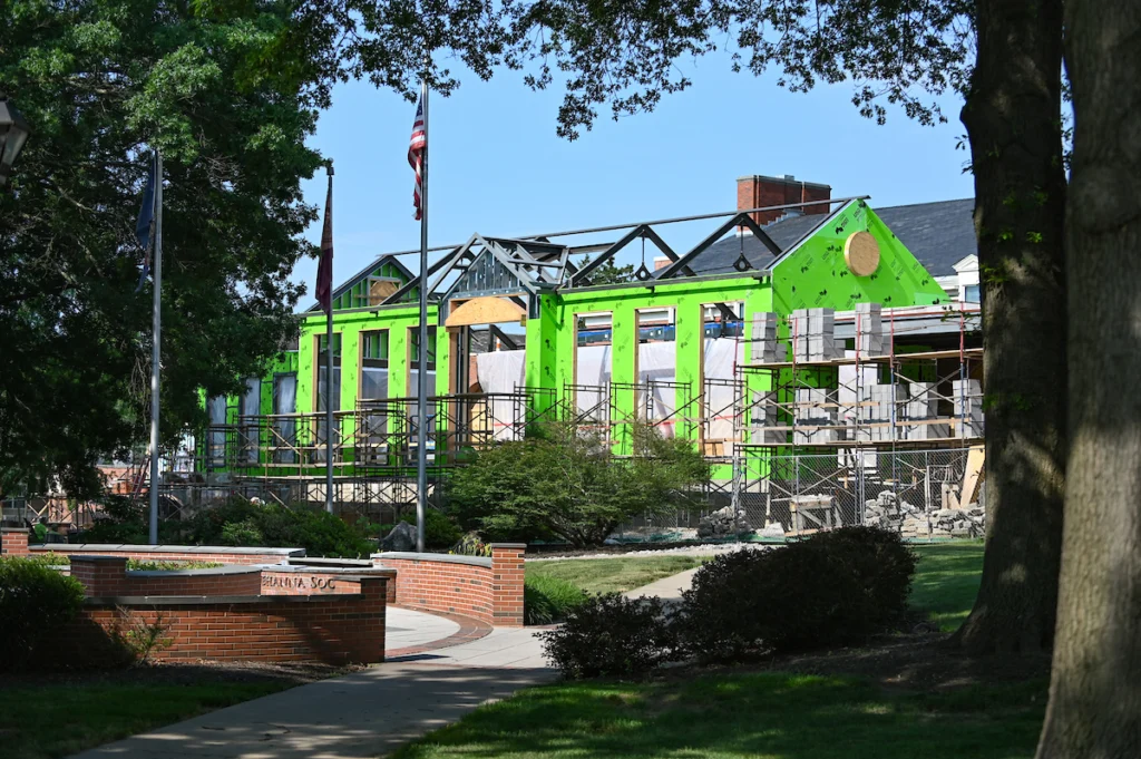A partially constructed building with green sheathing and scaffolding is surrounded by trees and a walkway; an American flag is visible in the background.