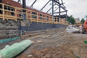 Construction site with partially built brick building, exposed steel framework, temporary wooden railings, and construction materials scattered on the ground.