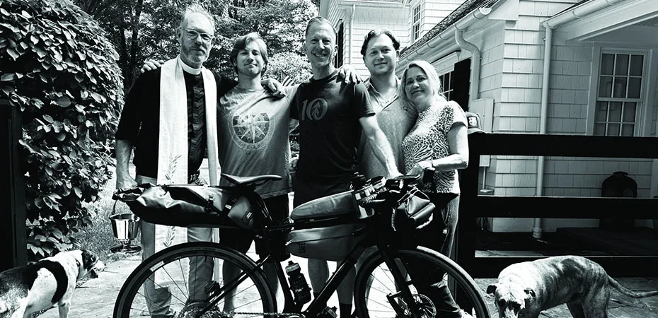 Five people stand together outdoors behind a loaded bicycle, with two dogs nearby and a house in the background.