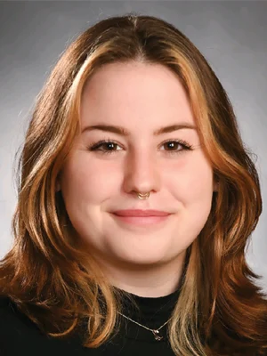 A young woman with light brown hair, a subtle smile, and a gold septum ring, wearing a black top and necklace, poses in front of a neutral background.