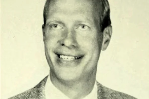 A black-and-white portrait of a man in a suit jacket and tie, smiling, facing the camera against a plain background.