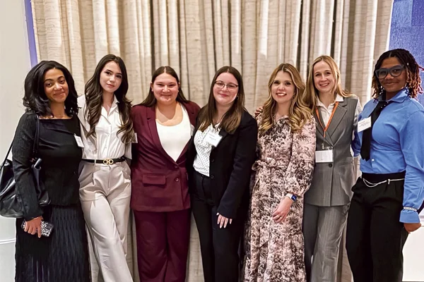 Seven women in business attire stand side by side indoors, smiling at the camera in front of beige curtains.