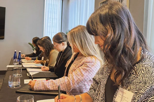 Six women sit side by side at a conference table, writing on documents with pens, with papers, water glasses, and bottles arranged in front of them.