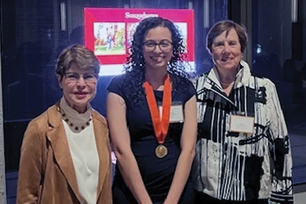 Three women stand indoors; the woman in the center wears a medal around her neck. A brightly lit screen with unclear content is visible in the background.