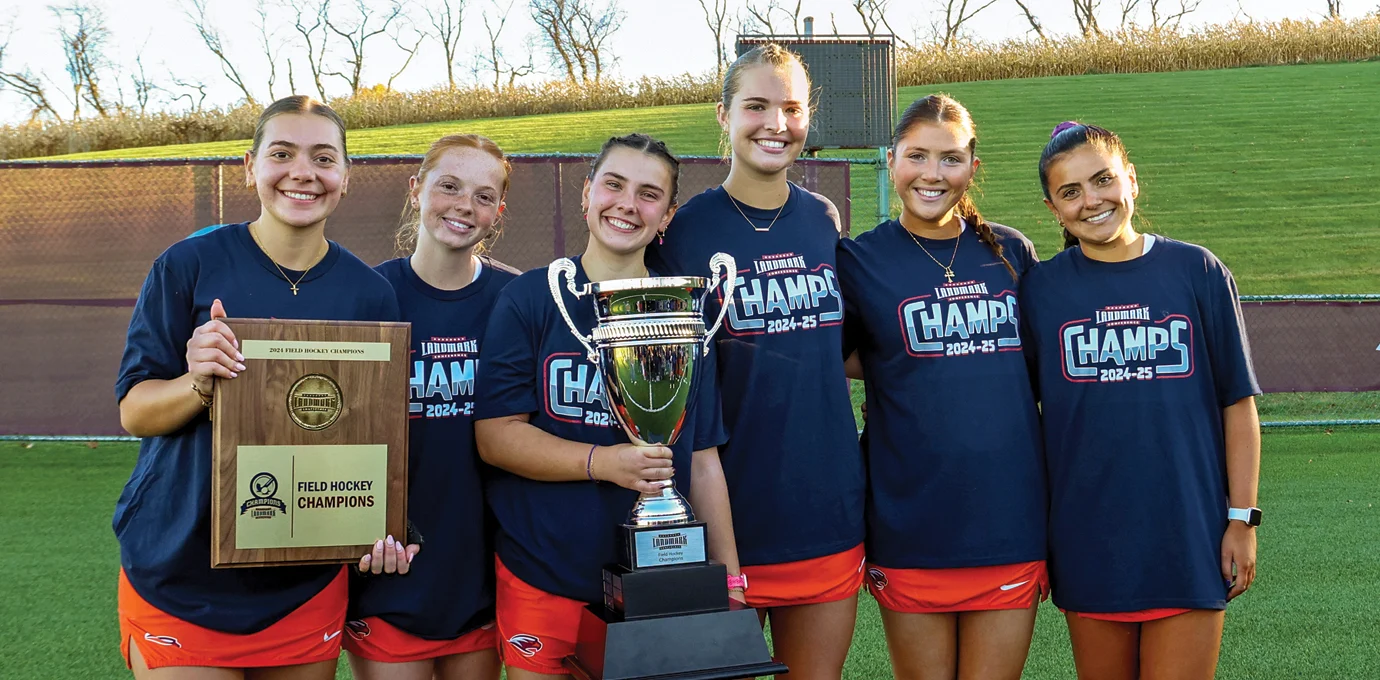 Six young women in matching shirts stand on a field holding a championship trophy and plaque, smiling at the camera.
