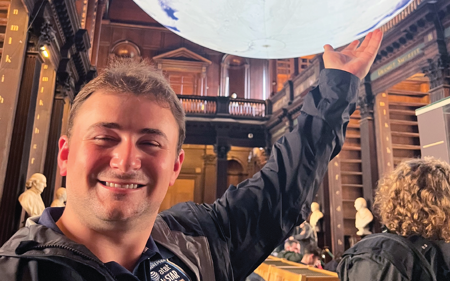 Person smiling and posing with a raised hand under a large sphere in a library-like setting with wooden shelves and busts.
