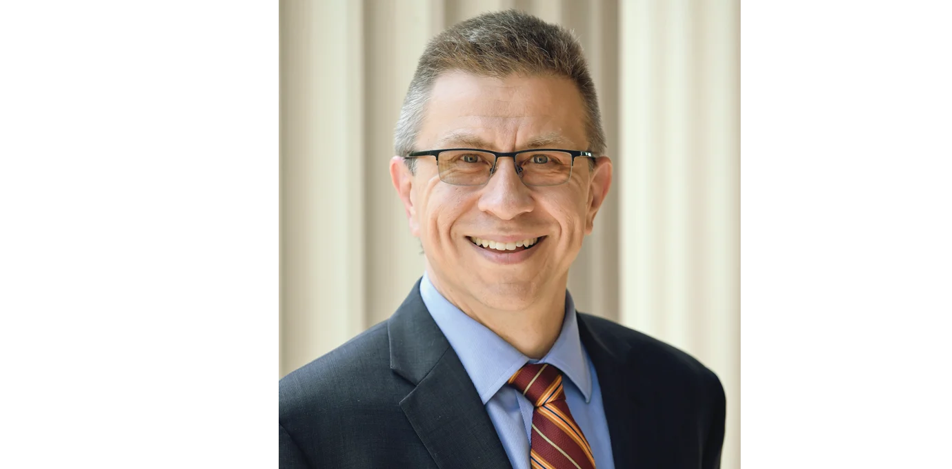 A man wearing glasses, a dark suit, blue shirt, and striped tie stands in front of a light-colored background, smiling at the camera.