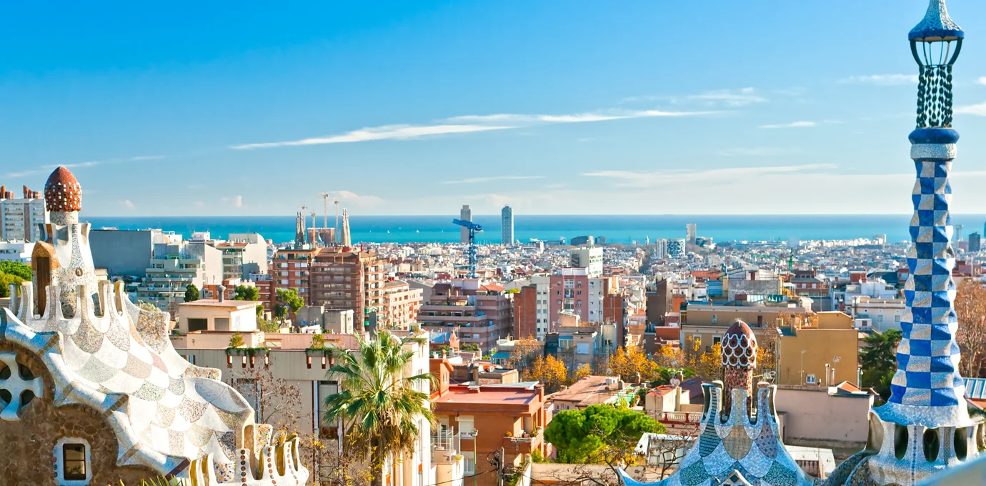 Panoramic view of Barcelona city with distinctive mosaic architecture in the foreground and the Mediterranean Sea in the background under a clear blue sky.