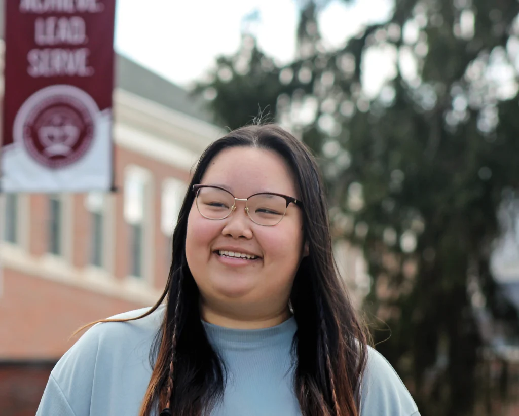 A person with long dark hair and glasses smiles outdoors in front of a blurred building and a maroon banner.
