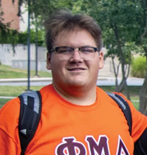 A person wearing glasses and an orange shirt with Greek letters stands outdoors, carrying a backpack. Trees and a building are visible in the background.