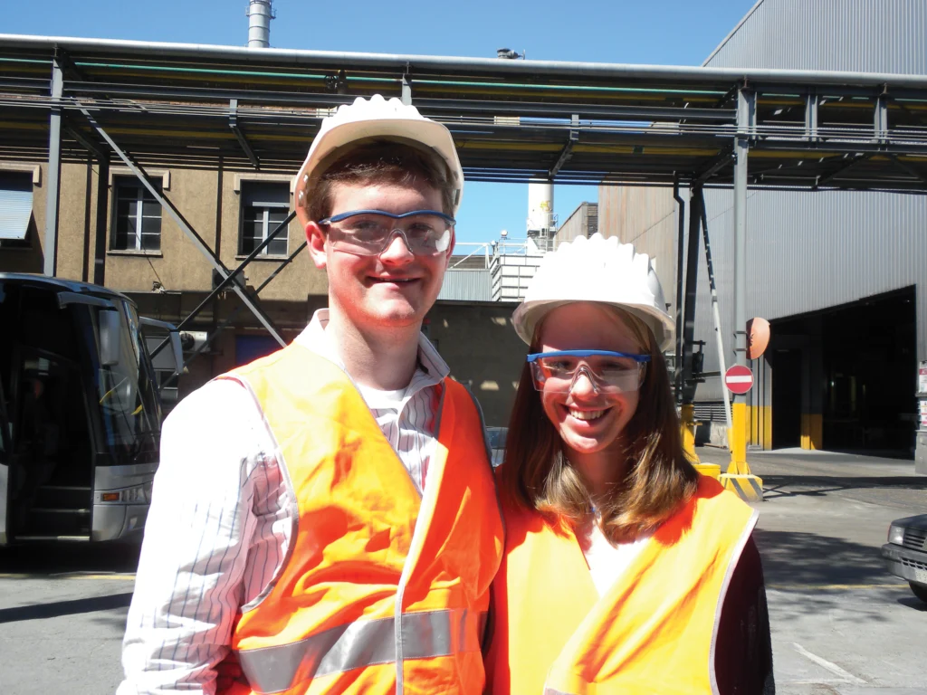 Two people wearing orange safety vests, hard hats, and safety glasses stand outdoors at an industrial facility.