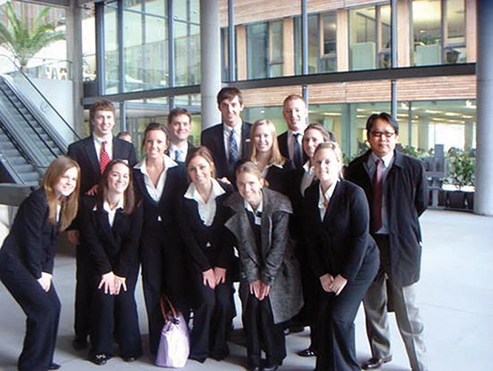 A group of thirteen people in business attire pose for a photo inside a modern office building with glass walls and visible staircases.