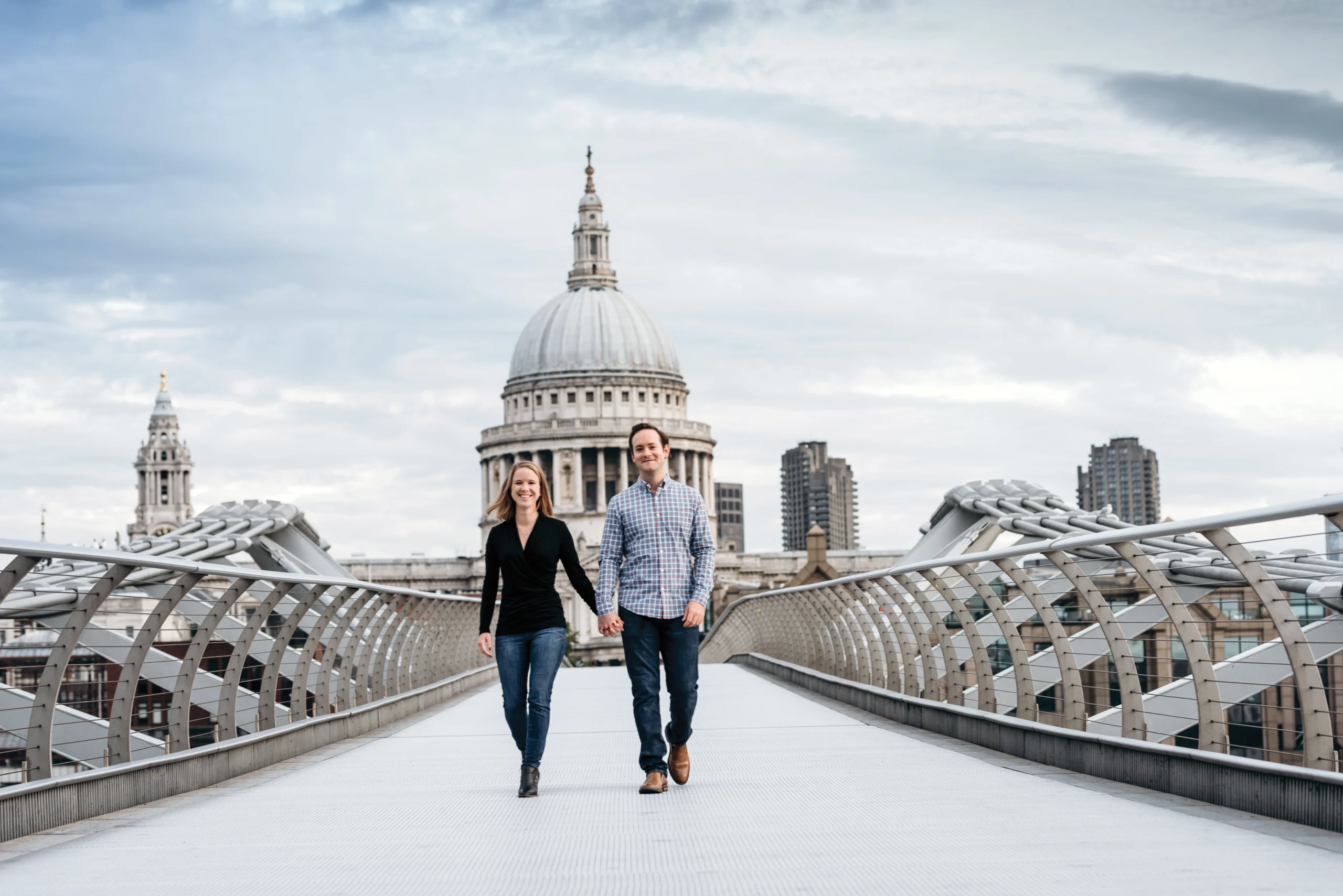 A couple walks hand in hand on the Millennium Bridge in London, with St. Paul’s Cathedral and city buildings visible in the background under a cloudy sky.