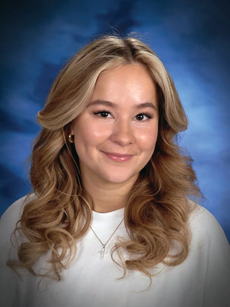 A young woman with long, wavy blonde hair is smiling at the camera, wearing a white top and a cross necklace, against a blue studio background.