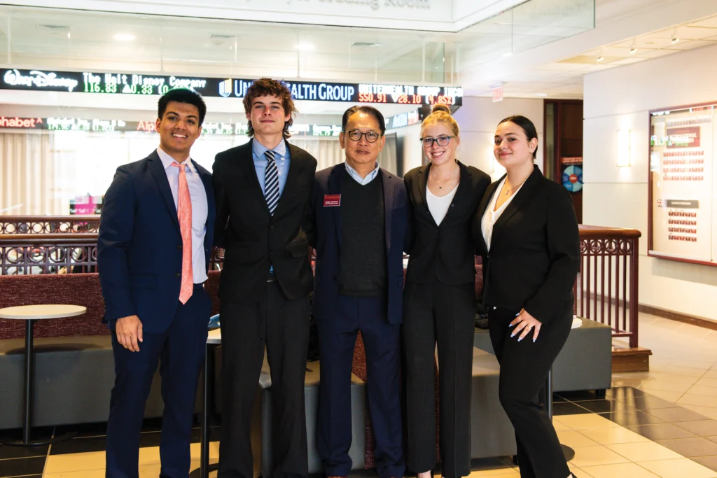 Five people in business attire stand together and pose for a group photo in an office lobby with an electronic stock ticker in the background.