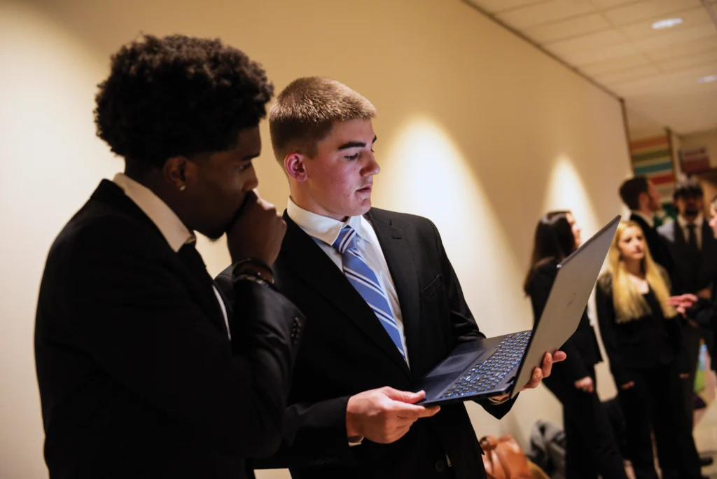 Two men in suits stand indoors, one holding and showing something on a laptop to the other, with several people in business attire in the background.