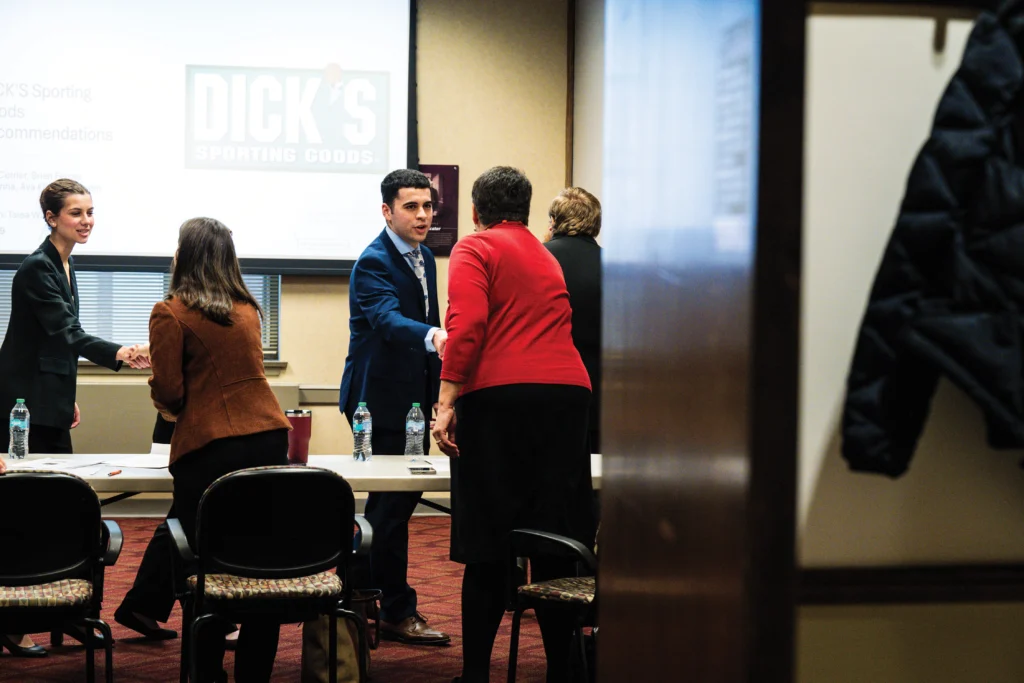 People in business attire shake hands and converse in a conference room, with a Dick's Sporting Goods presentation displayed in the background.
