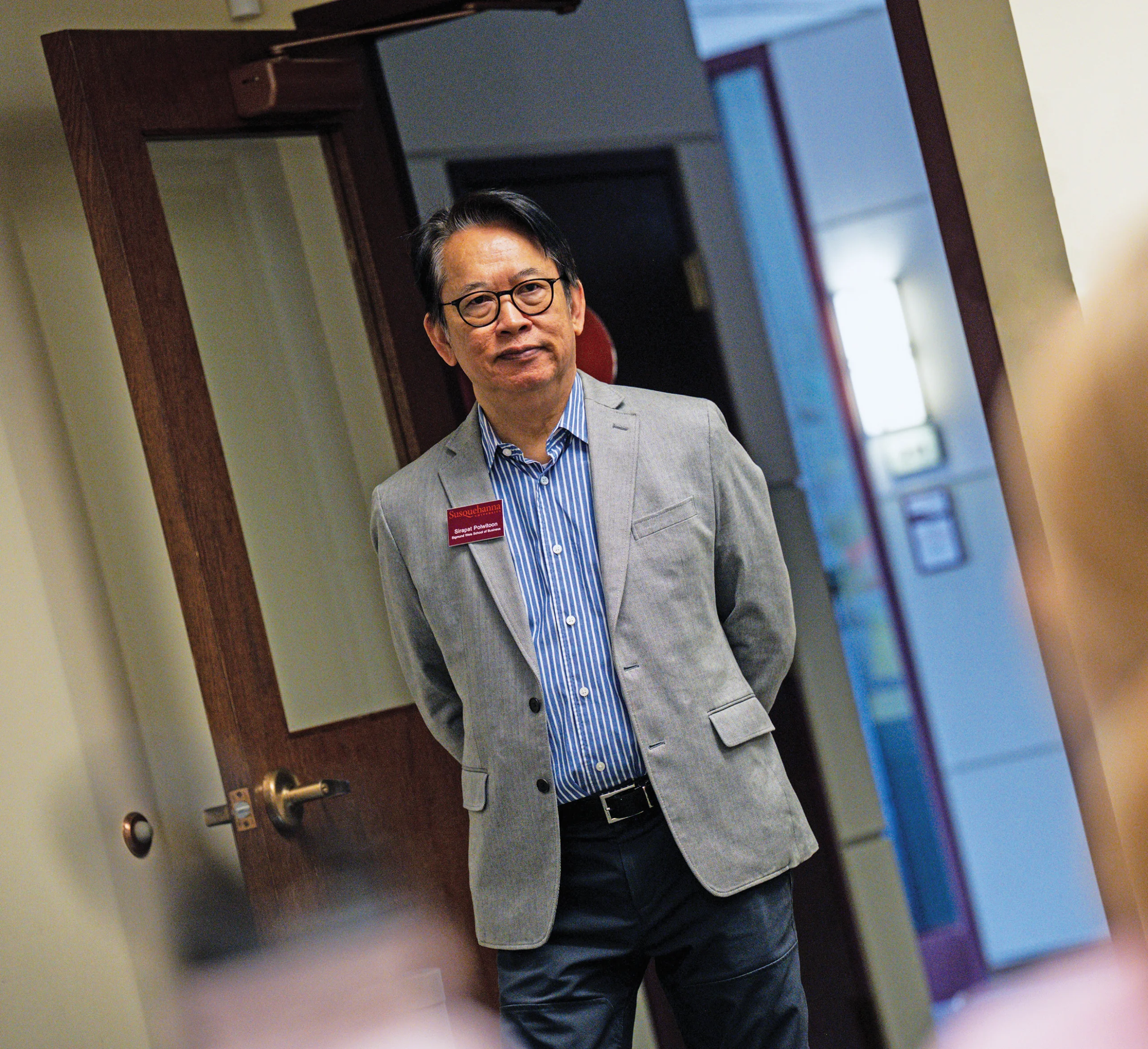 A man in a gray blazer and striped shirt stands near an open door in an office or classroom setting, looking forward with his hands behind his back.