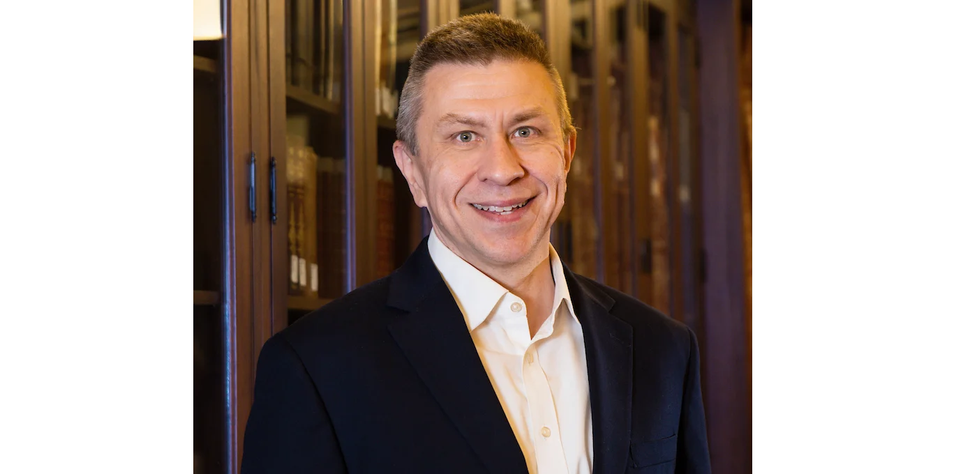 A man in a dark suit and white shirt stands in front of a bookshelf, smiling at the camera.