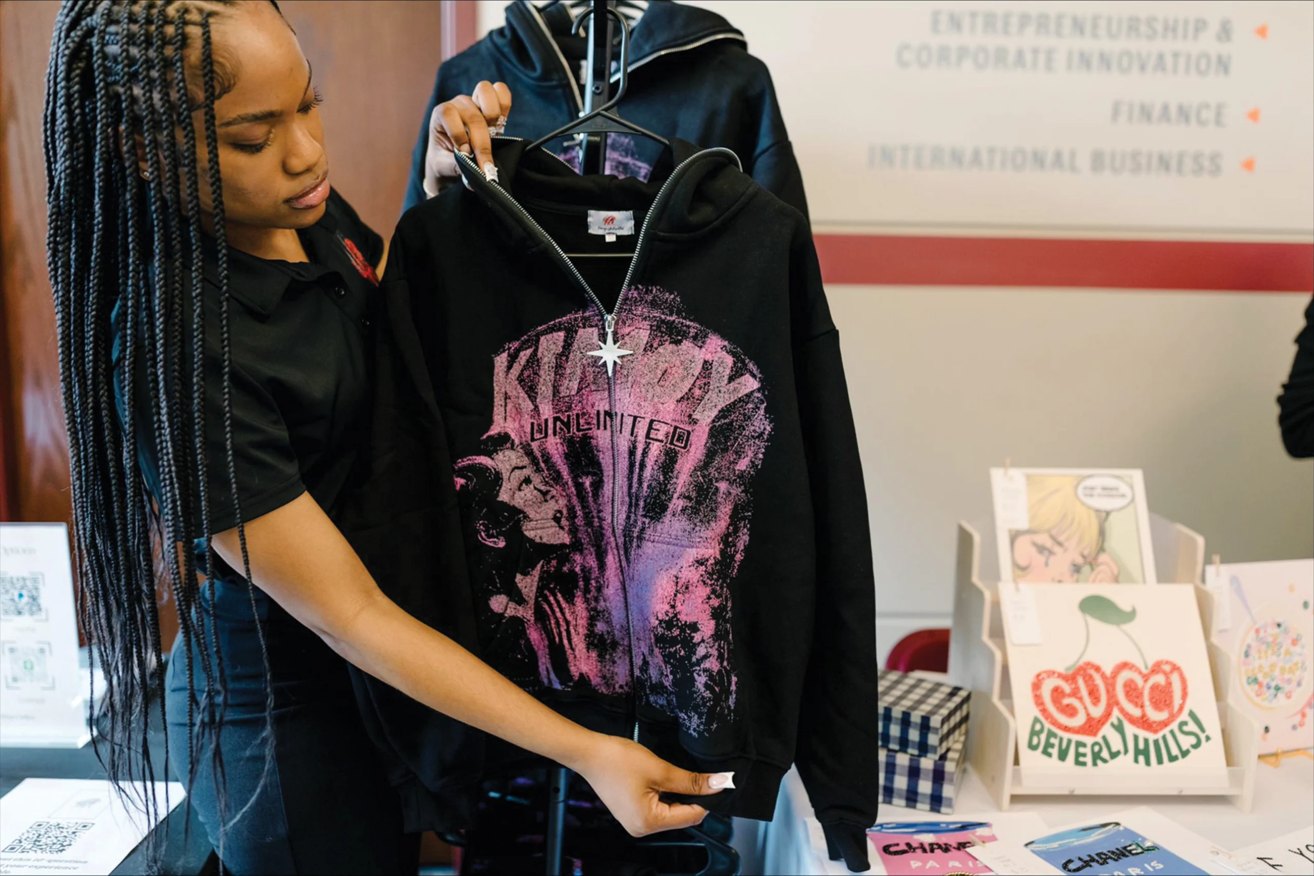A woman holds up a black hoodie with a purple graphic design at a display table with business-related signs and merchandise.