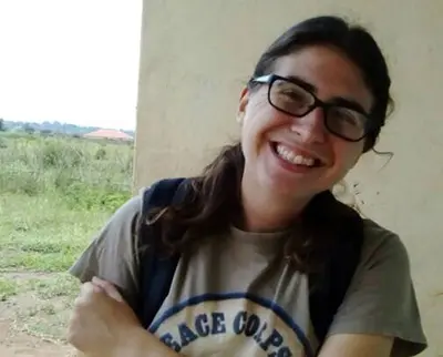 A person with long dark hair and glasses is smiling, wearing a Peace Corps T-shirt. They stand outdoors near a building with a grassy field and distant trees in the background.