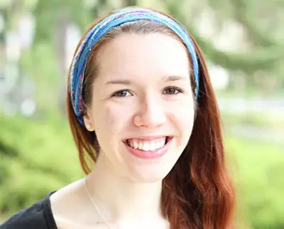 A woman with long red hair smiles at the camera. She wears a colorful headband and a black top. The background is a blurred outdoor setting with greenery.