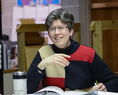 A person with short hair and glasses is smiling while sitting at a table. They are wearing a multicolored patchwork sweater and have a pen and notebook in front of them. A travel mug is also visible. The background includes bookshelves and papers.