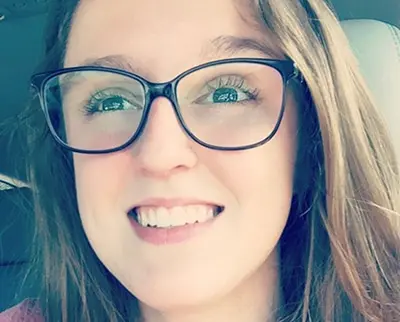 A person with long hair and glasses is smiling while sitting in a car. They have a natural expression and are looking slightly off to the side. The background is blurred, suggesting they are inside the vehicle.