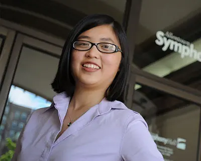 A woman with glasses and shoulder-length black hair smiles, wearing a lavender blouse. She stands in front of glass doors with the word “Symphony” partially visible.