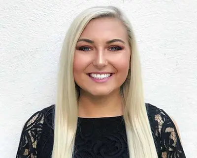 Smiling person with long blonde hair, wearing a black lace top, standing against a plain white background.