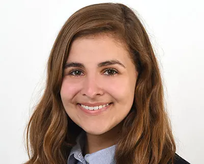 A person with long brown hair smiling, wearing a light-colored shirt and a dark jacket against a plain background.