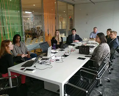 A group of eight people sit around a conference table in a modern office. Laptops and documents are on the table as they engage in discussion. The room has glass walls with colorful decorations visible outside.