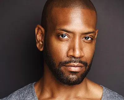 Portrait of a man with a shaved head and beard, wearing a gray shirt against a dark background. His expression is neutral and he is looking directly at the camera.