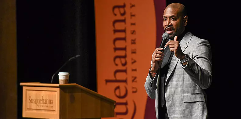 A man in a gray suit speaks into a microphone at a podium with a coffee cup, in front of a 体育买球 University banner.