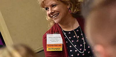 A smiling woman with curly blonde hair wears a lanyard with name tags reading 