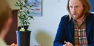 A man with long red hair and a beard, wearing a plaid shirt and navy cardigan, sits at a desk having a conversation. A potted plant and papers are on the table in front of him.
