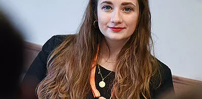 A woman with long brown hair, wearing a black top, a gold necklace, and an orange lanyard, looks at the camera and smiles softly. The background is out of focus.