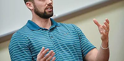 A man wearing a blue striped polo shirt gestures with both hands while speaking, standing in front of a blurred classroom setting.