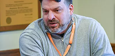 A middle-aged man with short dark hair and a beard, wearing a light gray zip-up sweater and an orange lanyard, sits indoors and appears to be speaking or listening attentively. A blurred plaque is visible in the background.