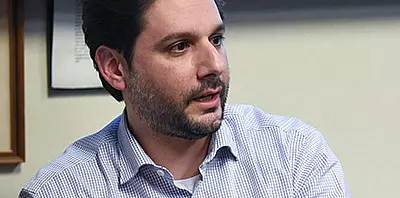 A man with short dark hair and a trimmed beard, wearing a light-colored checkered shirt, sits indoors and looks to his left, appearing to be engaged in conversation.