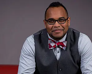 A person wearing glasses, a gray vest, a light blue shirt, and a red and white checkered bow tie is smiling at the camera against a gray background.