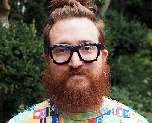 A person with a long red beard and mustache, wearing large black glasses and a colorful shirt, stands in front of a blurred green foliage background.