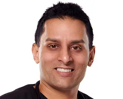 A person with short dark hair and a friendly smile is facing the camera. They are wearing a black shirt and a stethoscope around their neck against a plain white background.