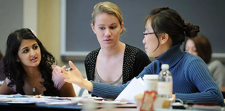 Three women sit at a table covered with papers and cups, engaged in a focused discussion in a classroom setting. One woman gestures as she speaks while the others listen attentively.
