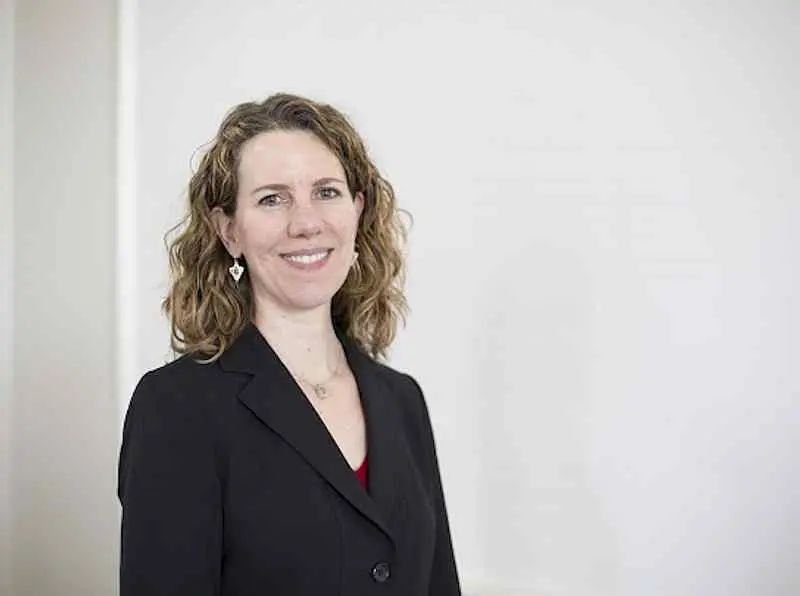 Woman with curly brown hair wearing a black blazer and red top stands smiling in front of a plain white background.
