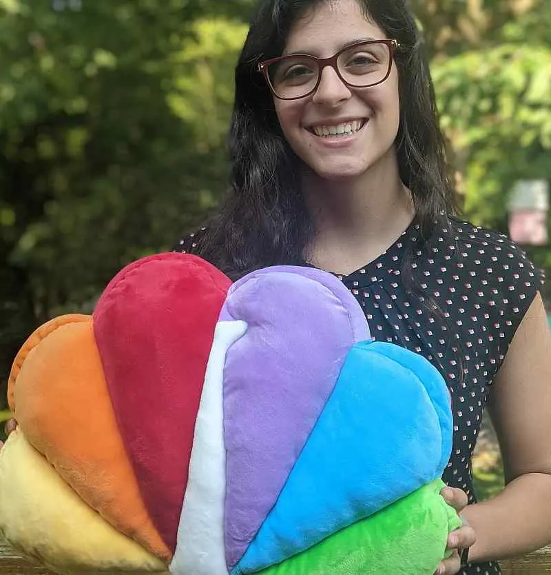 Smiling person with long dark hair and glasses holding a plush pillow shaped like a colorful rainbow logo, featuring red, orange, yellow, green, light blue, and purple sections. The background shows greenery.