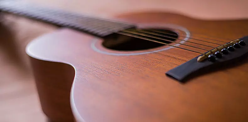 Close-up view of an acoustic guitar showing the wooden body, strings, bridge, and part of the fretboard, with a shallow depth of field creating a soft, blurred background.