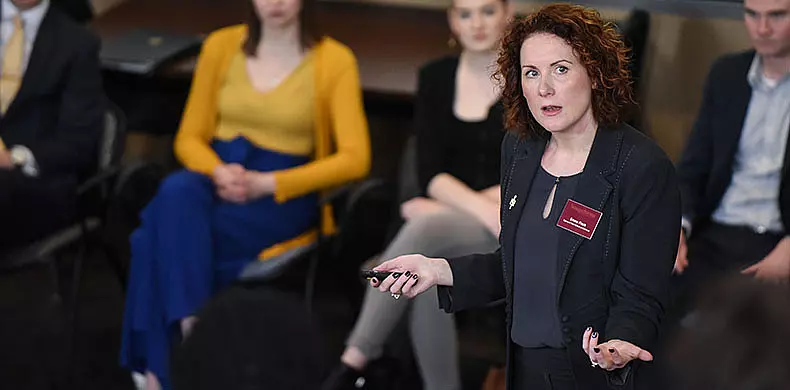 A woman with curly red hair, wearing a dark blazer and holding a remote, speaks to a seated audience. Several people, both men and women, are visible in the background listening attentively.
