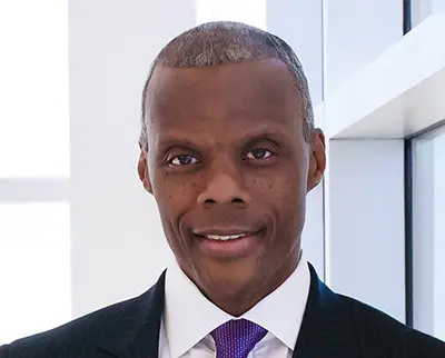 A person wearing a dark striped suit, purple tie, and white shirt smiles in a bright indoor setting. The background features large windows with white frames.
